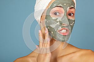 Beautiful cheerful teen girl applying facial clay mask. Beauty treatments, isolated on blue background