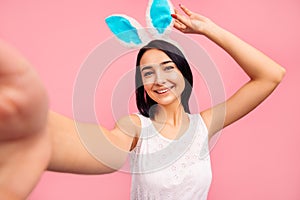 Beautiful cheerful woman in rabbit ears makes a selfie, video call, easter, in the studio on a pink background