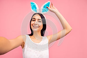 Beautiful cheerful woman in rabbit ears makes a selfie, video call, easter, in the studio on a pink background