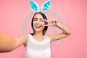 Beautiful cheerful woman in rabbit ears takes a selfie, shows two fingers, victory sign, video call, easter