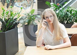 Portrait of a cheerful young caucasian blond business woman sitting on the table, talking at phone and looking at camera. Concept