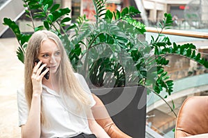 Portrait of a cheerful young caucasian blond business woman sitting on the table, talking at phone and smiling. Surprised face.