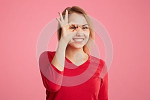 Beautiful cheerful young female makes ok sign, gestures in pink studio, shows her approval and like, dressed in red casual sweater