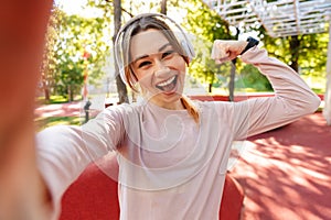 Beautiful cheerful young fitness sports woman posing outdoors in park listening music with earphones take selfie by camera