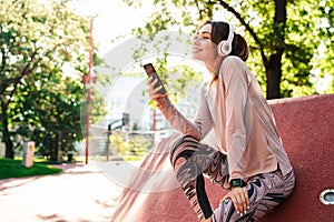 Beautiful cheerful young fitness sports woman posing outdoors in park listening music with earphones using mobile phone
