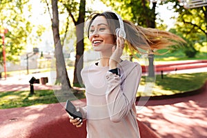 Beautiful cheerful young fitness sports woman posing outdoors in park listening music with earphones using mobile phone