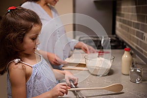 Beautiful child, adorable little girl standing at kitchen counter top, holding a wooden spoon on the blurred background of her mom