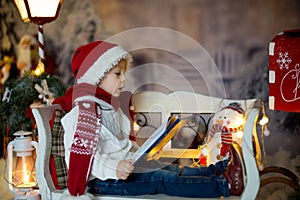 Beautiful child, blond toddler boy, reading a book, sitting on a bench with christmas decoration