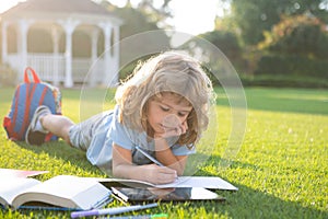 Beautiful child boy with book writing notes in copybook on grass background. Kids reading book in park.