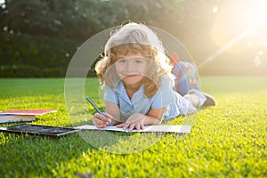 Beautiful child boy writing notes in copybook, laying on grass on the meadow background. Kids reading book in park.