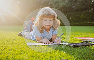 Beautiful child boy writing notes in copybook, laying on grass on the meadow background. Kids reading book in park.