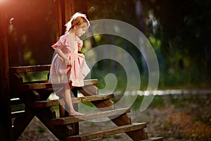 Portrait of little Caucasian girl wearing pink dress standing on wooden bridge, covering face and crying in forest