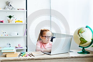 beautiful child in eyeglasses using laptop while studying at desk photo