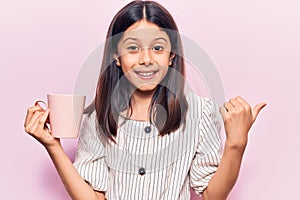 Beautiful child girl holding coffee pointing thumb up to the side smiling happy with open mouth