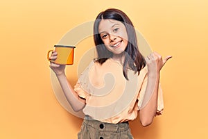 Beautiful child girl holding coffee pointing thumb up to the side smiling happy with open mouth