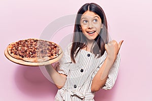 Beautiful child girl holding italian pizza pointing thumb up to the side smiling happy with open mouth