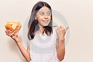 Beautiful child girl holding potato chip pointing thumb up to the side smiling happy with open mouth