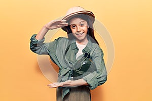 Beautiful child girl wearing explorer hat gesturing with hands showing big and large size sign, measure symbol