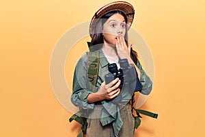 Beautiful child girl wearing explorer hat holding binoculars covering mouth with hand, shocked and afraid for mistake