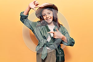 Beautiful child girl wearing explorer hat smiling making frame with hands and fingers with happy face