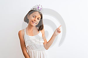 Beautiful child girl wearing princess crown standing over isolated white background with a big smile on face, pointing with hand