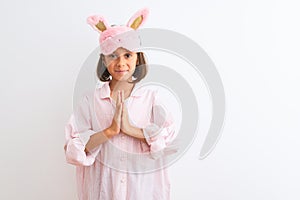 Beautiful child girl wearing sleep mask and pajama standing over isolated white background praying with hands together asking for