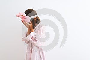 Beautiful child girl wearing sleep mask and pajama standing over isolated white background with sad expression covering face with