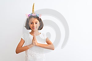 Beautiful child girl wearing unicorn diadem standing over isolated white background praying with hands together asking for
