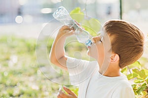 A beautiful child sitting on the grass drinks water from a bottle in the summer at sunset. Boy quenches his thirst on a hot day