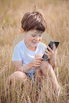 A beautiful child sitting on the grass speaks by phone in the summer at sunset. Boy communicates on mobile