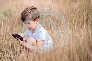 A beautiful child sitting on the grass speaks by phone in the summer at sunset. Boy communicates on mobile