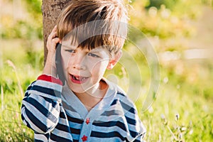 A beautiful child sitting on the grass speaks by phone in the summer at sunset. Boy communicates on mobile