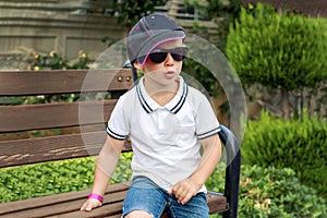 A beautiful child in sunglasses, a cap and a white polo shirt - sits on a bench and looks into the distance, thinking.