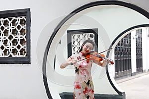 Beautiful Asian Chinese woman in traditional chi-pao cheongsam play violin in in a garden stand under arched door