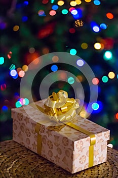 Beautiful Christmas gift with a gold bow under the tree against a background of multi-colored bokeh lights blurry out of focus