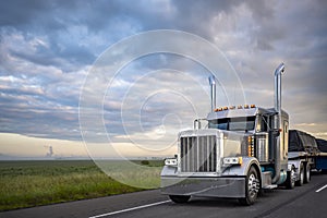 Beautiful classic big rig American semi truck with light transporting covered cargo on flat bed semi trailer driving on the