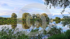 A beautiful cloudy sky in the evening over the river. The branches of trees and bushes lean over the water with ripples in which t