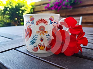 A beautiful coffee cup in hand-painted handmade folk patterns; red blooming geranium flower next to the cup set on a wooden table.