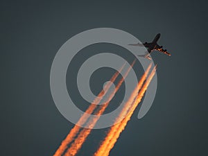 An airplane with contrails is illuminated by the last light of the setting sun