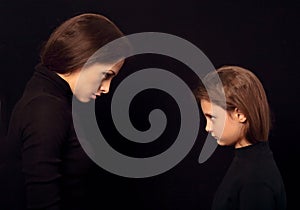 Beautiful concentrated angry mother and her seroius emotional daughter standing and looking each other on black shadow background