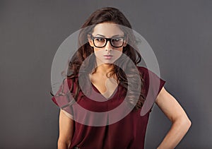 Beautiful concentrated serious brunette business woman looking in eye glasses with curly hair style in red silk blouse on grey