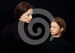 Beautiful concentrated serious mother and her angry emotional thinking daughter looking each other on black shadow background.