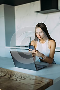 Beautiful concentrated young woman typing text message on mobile phone while calculating bills in kitchen, sitting at table with