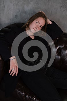 beautiful confident brunette woman in black on a brown leather couch in grey dark room. business woman. feminine