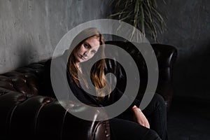 beautiful confident brunette woman in black on a brown leather couch in grey dark room. business woman. feminine