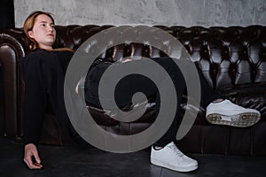 beautiful confident brunette woman in black on a brown leather couch in grey dark room. business woman. feminine