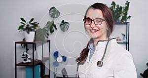 Beautiful confident Caucasian female healthcare professional in lab coat turns head at camera posing at light office.