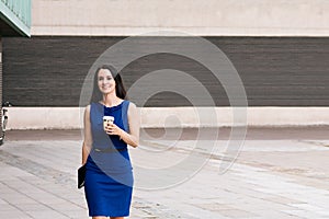 Beautiful confident Hispanic businesswoman with smile walking outdoors near office building with coffee in hand