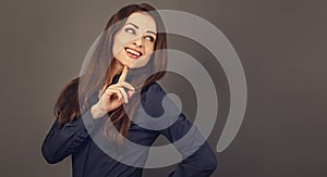 Beautiful confident thinking toothy smiling business woman with finger under the face in blue shirt on grey background with empty
