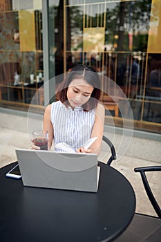 Beautiful confident woman in casual clothing reading book and drinking coffee while sitting in cafe with laptop and notebook